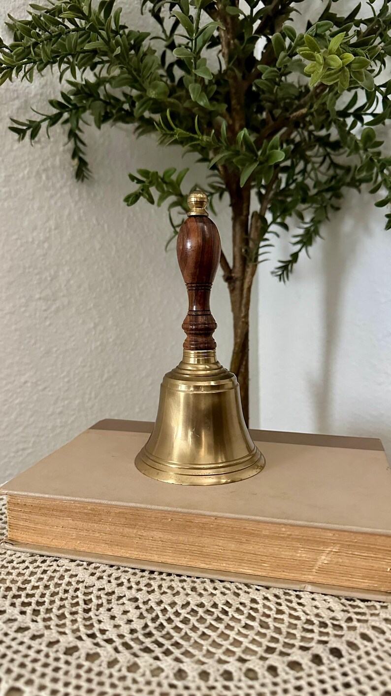 May include: A hand bell featuring a polished brass bell base and a dark brown wooden handle with a small brass finial at the top. The handle has a turned, ribbed design, and the brass bell has a smooth, flared rim and a reflective metallic finish.