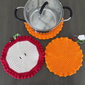 Puede incluir: Una olla de acero inoxidable con asa negra descansa sobre un salvamanteles de ganchillo naranja. Se ven dos salvamanteles de ganchillo adicionales: uno rojo y blanco con diseño de manzana y otro naranja liso. Todos los salvamanteles tienen un borde festoneado.