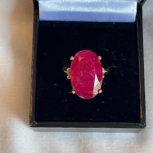May include: A ring featuring a large, oval-cut, deep red gemstone set in a gold-toned metal band with four prongs, presented inside a black jewelry box with a black velvet interior lining and a white satin trim along the inner lid edge.