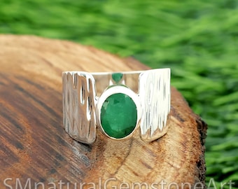 Puede incluir: Un anillo de plata con una gran esmeralda ovalada. El anillo tiene una banda texturizada y está sobre una superficie de madera con follaje verde de fondo. El texto "SMnaturalGemstoneArt" es visible.