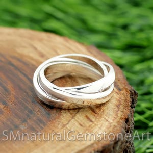 May include: A silver ring with three interlocking bands. The ring is made of polished metal and sits on a wooden surface. The ring is a piece of jewelry.