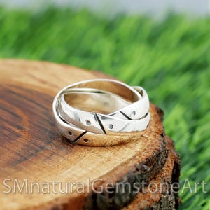 Puede incluir: Un anillo de plata con un diseño entrelazado, con patrones geométricos y pequeños acentos negros. El anillo se muestra sobre una superficie de madera con el texto "SMnaturalGemstoneArt" visible.