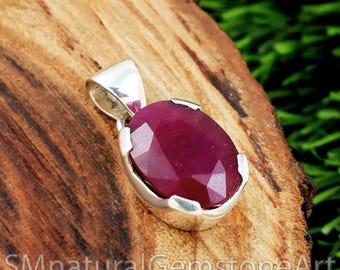 May include: A silver pendant with a large, faceted, oval-shaped ruby gemstone. The ruby is a deep red color and set in a polished silver bezel. The pendant is displayed on a wooden surface. The text "SMnaturalGemstoneArt" is visible at the bottom.