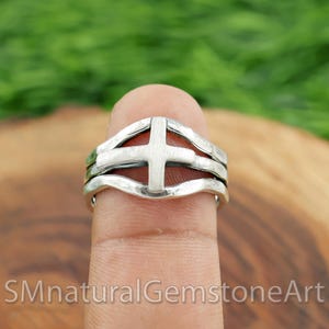 Peut inclure: Une bague en argent avec un motif de croix. La bague présente une bande superposée avec une croix en relief au centre. La bague est présentée sur un doigt sur un fond vert et marron flou.