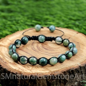 Puede incluir: Un brazalete de piedra de ágata verde con cordón y nudos negros. El brazalete está hecho con cuentas redondas y se muestra sobre una superficie de madera.