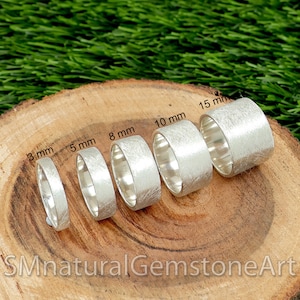 Könnte beinhalten: Sechs silberne Ringe in verschiedenen Größen von 3 mm bis 15 mm Durchmesser werden auf einer Holzoberfläche gezeigt. Die Ringe sind alle glatt und haben eine gebürstete Oberfläche. Die Ringe sind mit ihrer Größe in Millimetern beschriftet.