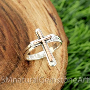 May include: A silver ring with a copper cross design. The ring is on a wooden surface.