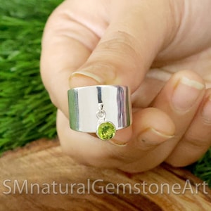 Op de afbeelding: Een zilveren ring met een groene edelsteen hanger. De ring is breed en heeft een plat oppervlak. De edelsteen is rond en heeft een lichtgroene kleur.
