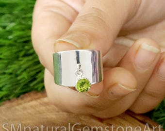 May include: A silver ring with a green gemstone pendant. The ring is wide and has a flat surface. The gemstone is round and has a light green color.