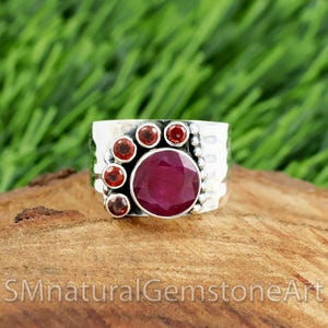 May include: A silver ring with a large, round, deep red ruby center stone, surrounded by smaller, round, red garnet stones. The ring band has a textured, hammered appearance. The text "SMnaturalGemstoneArt" is at the bottom.