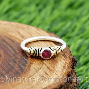 Puede incluir: Un anillo de plata con una banda dorada y una piedra preciosa redonda de color rojo intenso. El anillo está colocado sobre un trozo de madera con hierba verde en el fondo. El texto "SMnaturalGemstoneArt" es visible en la parte inferior.