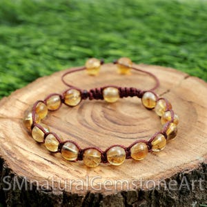 May include: A brown cord bracelet with a knotted closure and amber-colored beads. The bracelet is on a wooden surface.