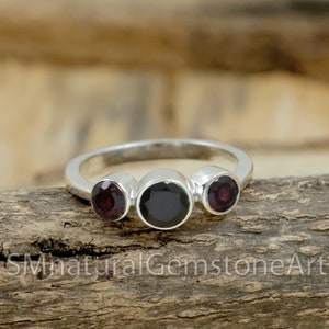 Puede incluir: Un anillo de plata con tres piedras redondas. La piedra central es negra y las dos piedras exteriores son moradas.