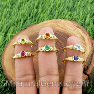 Może przedstawiać: Złote pierścionki z kolorowymi kamieniami szlachetnymi, w tym zielonymi, czerwonymi, żółtymi, niebieskimi i szmaragdowymi. Pierścionki są prezentowane na palcach na tle drewnianego tła. Pierścionki mają unikalny żebrowany wzór.