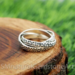 Puede incluir: Un anillo de plata con tres bandas entrelazadas. Cada banda tiene un patrón texturizado y punteado. El anillo se muestra sobre una superficie de madera con el texto "SMnaturalGemstoneArt" en la parte inferior.