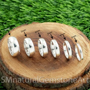 Puede incluir: Seis anillos de plata de diferentes anchos, de 3 mm a 8 mm, se muestran sobre una superficie de madera. Los anillos están dispuestos en fila, mostrando sus diferentes tamaños. Los anillos están muy pulidos y reflejan la luz.