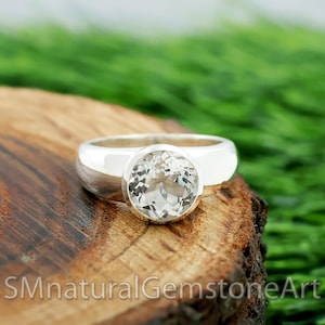 May include: A silver ring with a large, round, clear gemstone. The ring is set on a wooden surface with a green background. The text "SMnaturalGemstoneArt" is visible at the bottom.