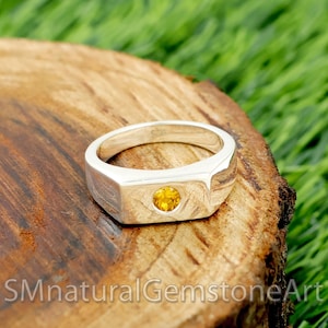 May include: A silver ring with a rectangular face and a round, yellow gemstone set in the center. The ring is displayed on a wooden surface with green grass in the background. The text "SMnaturalGemstoneArt" is at the bottom.