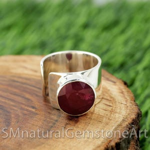 Puede incluir: Un anillo de plata con una gran piedra preciosa de rubí redonda facetada engastada en un engaste de bisel. El anillo es ancho y tiene un diseño simple y moderno.