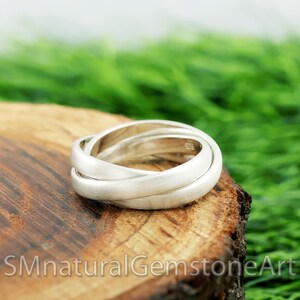 Può includere: Un anello d'argento con tre fasce intrecciate, appoggiato su una superficie di legno. L'anello ha una finitura opaca e un design semplice. Lo sfondo è sfocato, con fogliame verde.