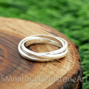 May include: A silver-colored ring with three interlocking bands. The ring is displayed on a piece of wood with a natural grain. The background is a blurred green. The text "SMnaturalGemstoneArt" is visible at the bottom.