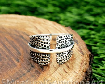 May include: A silver ring with a textured, dotted pattern on the band. The ring has a unique open design with a horizontal band crossing the textured sections. The ring is displayed on a wooden surface.
