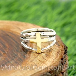 Peut inclure: Une bague en argent avec une croix dorée au centre. La bague comporte trois bandes et est posée sur une surface en bois avec un feuillage vert en arrière-plan. Le texte "SMnaturalGemstoneArt" est en bas.
