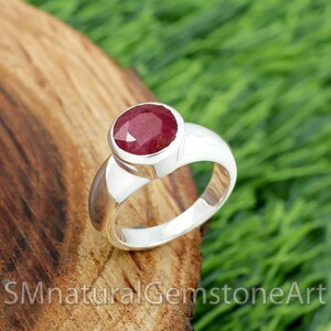 May include: A silver ring with a large, oval-cut ruby gemstone. The ruby is a deep red colour and set in a polished silver band. The ring is displayed on a wooden surface with green grass in the background. The text "SMnaturalGemstoneArt" is visible.