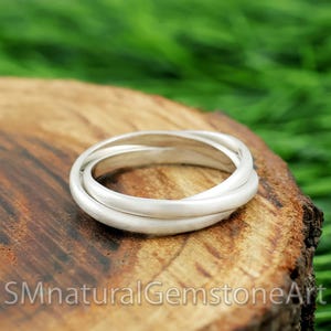 May include: A silver ring featuring three interlocking bands. The ring is set on a wooden surface, with the text "SMnaturalGemstoneArt" visible. The background is a blurred green.