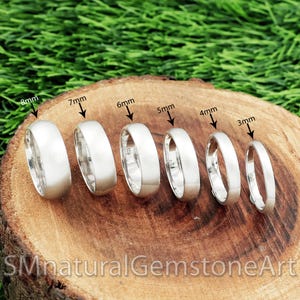 May include: A selection of silver rings, ranging from 3mm to 8mm in width, presented on a wooden surface. The rings are arranged by size, with each ring's width clearly indicated in millimetres. The background features blurred green foliage.