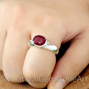 Könnte beinhalten: Ein silberner Ring mit einem großen, oval geschliffenen Rubin. Der Rubin hat eine tiefrote Farbe und ist in einem polierten Silberband gefasst. Der Ring wird an einem Finger gezeigt, um die Größe und Brillanz des Edelsteins hervorzuheben. Der Text "SMnaturalGemstoneArt" ist unten sichtbar.