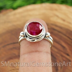 Puede incluir: Un anillo de plata con una piedra preciosa de rub&iacute; rojo engastada en un dise&ntilde;o de alambre envuelto. El anillo se lleva en un dedo.