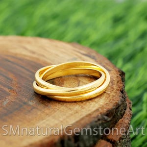 Könnte beinhalten: Ein goldfarbener, ineinandergreifender Ring, der auf einer Holzoberfläche liegt. Der Ring hat drei ineinander verschlungene Bänder, die eine durchgehende Schleife bilden. Der Hintergrund ist ein verschwommenes Grün, was auf eine natürliche Umgebung hindeutet. Der Text "SMnaturalGemstoneArt" ist unten sichtbar.