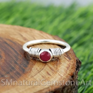 May include: A silver ring with a central, round, deep red gemstone. The band has a coiled wire design on either side of the gemstone. The ring is set against a wooden surface with a green background.