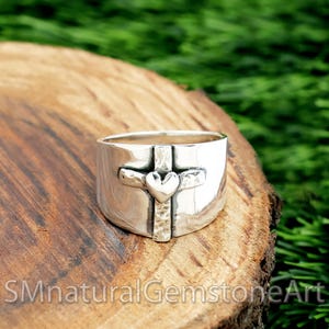 Puede incluir: Un anillo ancho de plata con un diseño de cruz y corazón. La cruz está centrada en el anillo y tiene un acabado texturizado. El corazón está en el centro de la cruz. El anillo está sobre una superficie de madera.