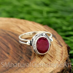 Könnte beinhalten: Ein silberner Ring mit einem tief roten ovalen Edelstein, der in einem gedrehten Drahtdesign gefasst ist. Der Ring hat zwei kleine silberne Perlen auf der Band.
