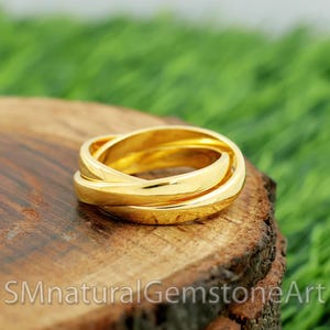 Pode incluir: Um anel dourado com três faixas entrelaçadas. O anel, feito de metal brilhante, está sobre uma superfície de madeira com um fundo verde desfocado. O texto "SMnaturalGemstoneArt" é visível. Uma joia.