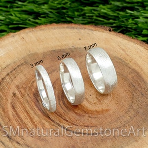 Op de afbeelding: Vier zilveren ringen in verschillende breedtes, 3 mm, 5 mm en 7 mm, gepresenteerd op een houten oppervlak. De ringen hebben een geborstelde afwerking.