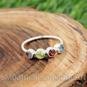 Könnte beinhalten: Ein silberner Ring mit fünf Edelsteinen in Lila, Grün, Rot und Blau. Der Ring liegt auf einer Holzoberfläche. Der Text "SMnaturalGemstoneArt" ist im unteren Teil des Bildes zu sehen.
