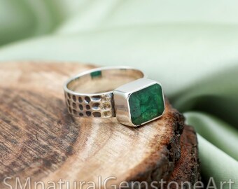 Puede incluir: Un anillo de plata con una esmeralda rectangular de color verde intenso engastada en un bisel liso. La banda del anillo tiene un acabado martillado texturizado con una serie de pequeñas hendiduras uniformes en la superficie exterior.