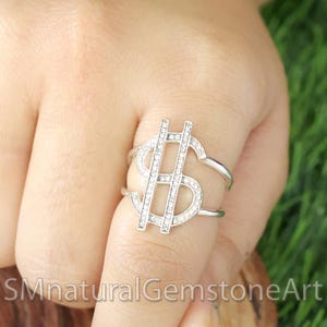 May include: A silver ring with a large, clear crystal dollar sign design.