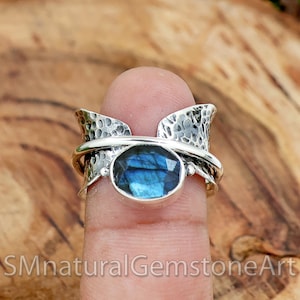 May include: A silver ring with a blue labradorite gemstone set in a hammered silver band. The ring has a unique design with two curved silver pieces that wrap around the gemstone.