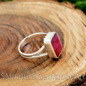 May include: A silver ring with a large, rectangular, faceted ruby gemstone. The ruby is a vibrant red color and set in a bezel setting. The ring band has a split shank design, adding a unique detail to the jewelry.