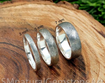 Könnte beinhalten: Vier silberne Ringe mit gebürsteter Oberfläche, auf einer Holzoberfläche präsentiert. Die Ringe sind mit ihrer Breite in Millimetern gekennzeichnet: 3 mm, 5 mm, 7 mm.