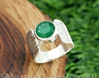 Puede incluir: Un anillo de plata con una gran gema redonda de color verde esmeralda. El anillo tiene un diseño único con detalles en forma de pluma a ambos lados de la gema. Está sobre una superficie de madera con follaje verde de fondo. El texto "SMnaturalGemstoneArt" es visible.