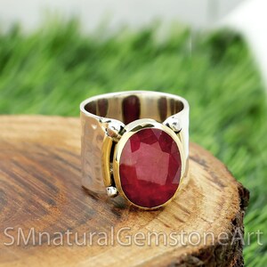 May include: A silver ring with a hammered finish and a large, oval-shaped ruby gemstone set in a gold bezel. The ring is on a wooden surface.