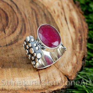 May include: A silver ring featuring a large, oval-shaped, faceted ruby gemstone. The ring band is textured with small, raised dots.