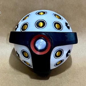 Puede incluir: Objeto esférico futurista con superficie blanca salpicada de detalles circulares dorados y negros. Una banda negra rodea el centro, y un detalle circular rojo y plateado está en la parte delantera. Tema mecánico o tecnológico.