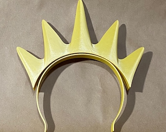 Puede incluir: Una diadema de color amarillo brillante con un accesorio rígido en forma de corona que presenta cinco puntas triangulares distintas orientadas hacia arriba. La superficie del material plástico muestra finas líneas de textura impresas en 3D.