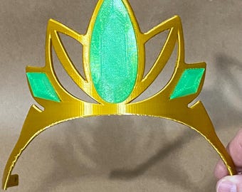 Puede incluir: Una corona dorada con detalles de gemas verdes. La corona tiene una parte superior puntiaguda y una base curva.